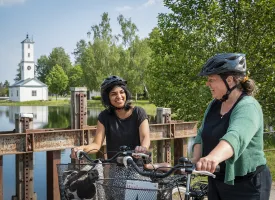 Zwei Frauen mit Fahrrädern stehen auf einer Brücke mit einer kleinen Kirche im Hintergrund.