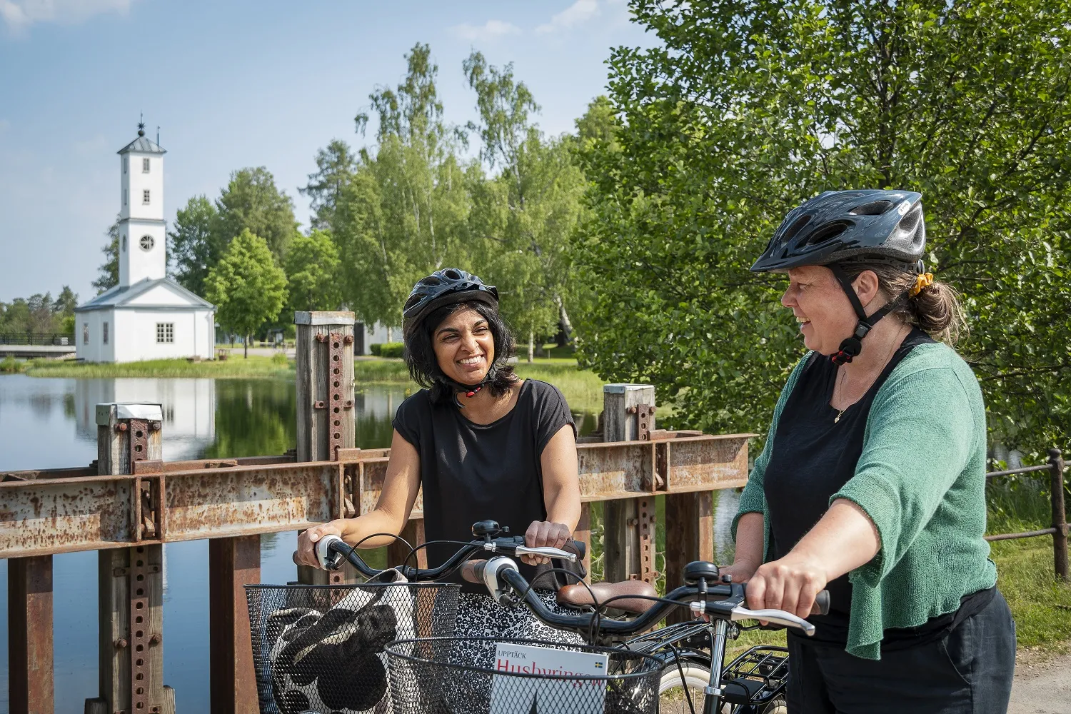 Twee vrouwen met fietsen staan op een brug met een kerkje op de achtergrond.