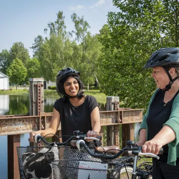 Zwei Frauen mit Fahrrädern stehen auf einer Brücke mit einer kleinen Kirche im Hintergrund.